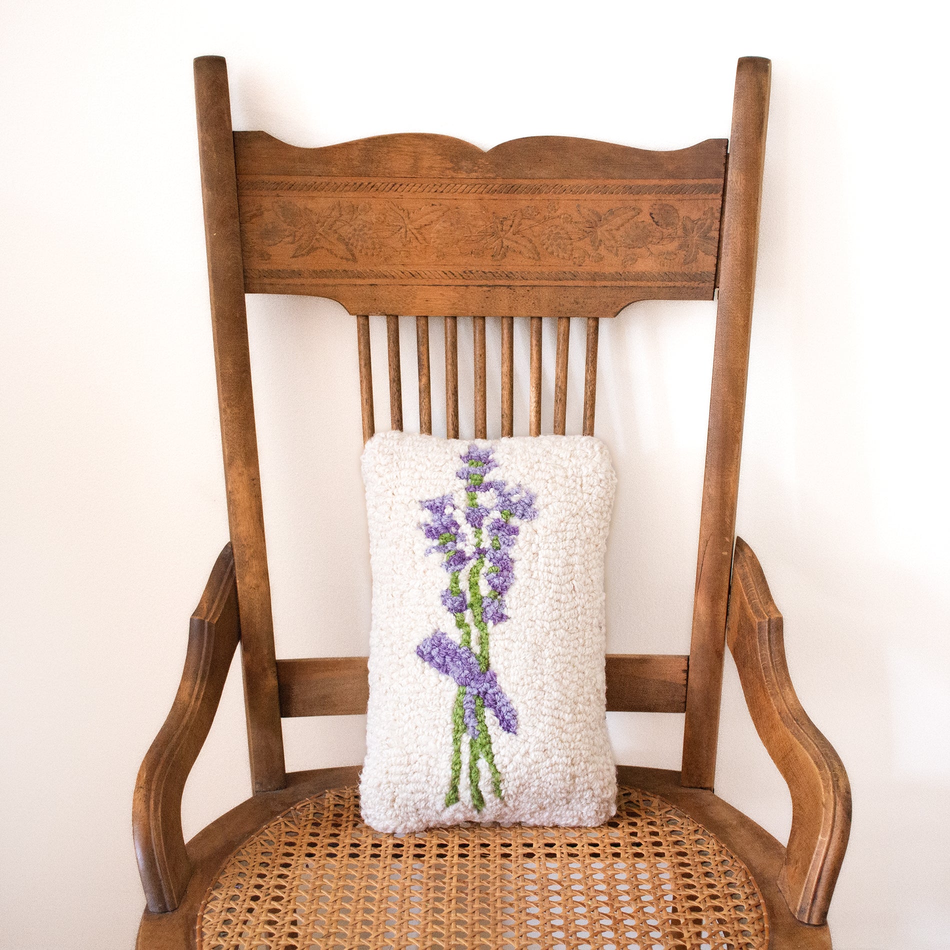 A Chandler 4 Corners brand Lavender Hooked Wool Handmade Pillow with a purple and green lavender design & a white background for the design, sitting on an antique wooden chair.