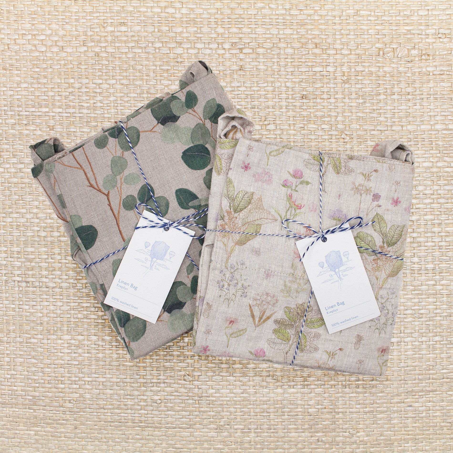 Two Linen Tales linen tote bags, folded and wrapped in blue and white striped twin. One bag has a eucalyptus design with a natural background, while the other has a pink and light green floral/leafy design with a natural background.